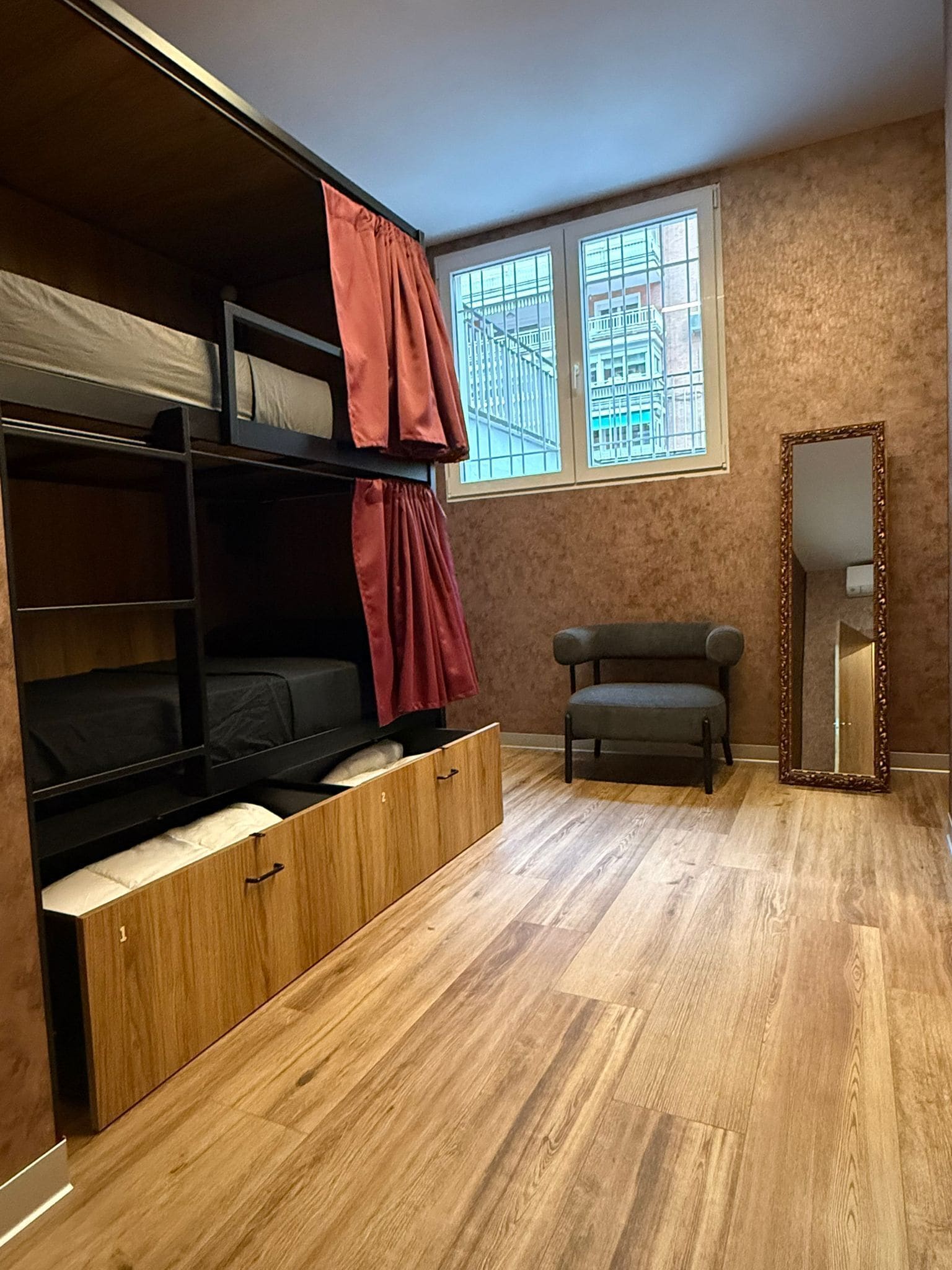A room with a bunk bed, a chair, a mirror, and a window with a view of buildings.