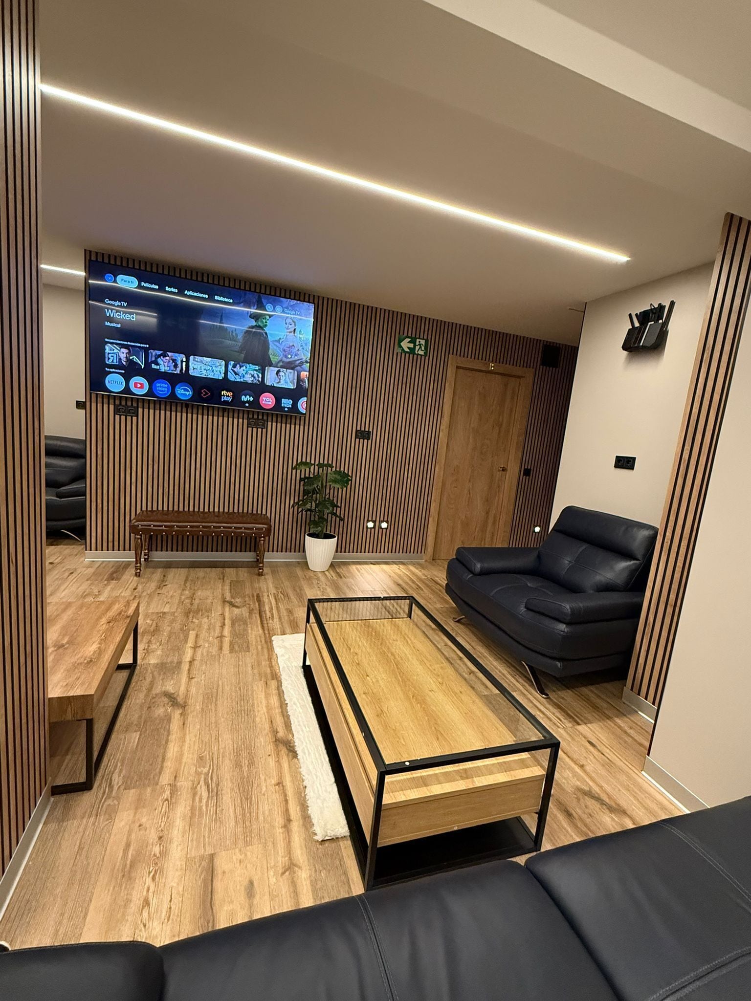 Un salon moderne avec un téléviseur à écran plat fixé au mur, un banc en bois, un canapé en cuir noir et une table basse.