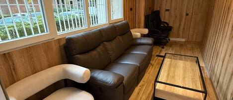 Un salon moderne avec un canapé en cuir marron, un fauteuil blanc et une petite table. De grandes fenêtres avec des barreaux en métal, une chaise de bureau noire et un plancher en bois complètent l’ensemble.