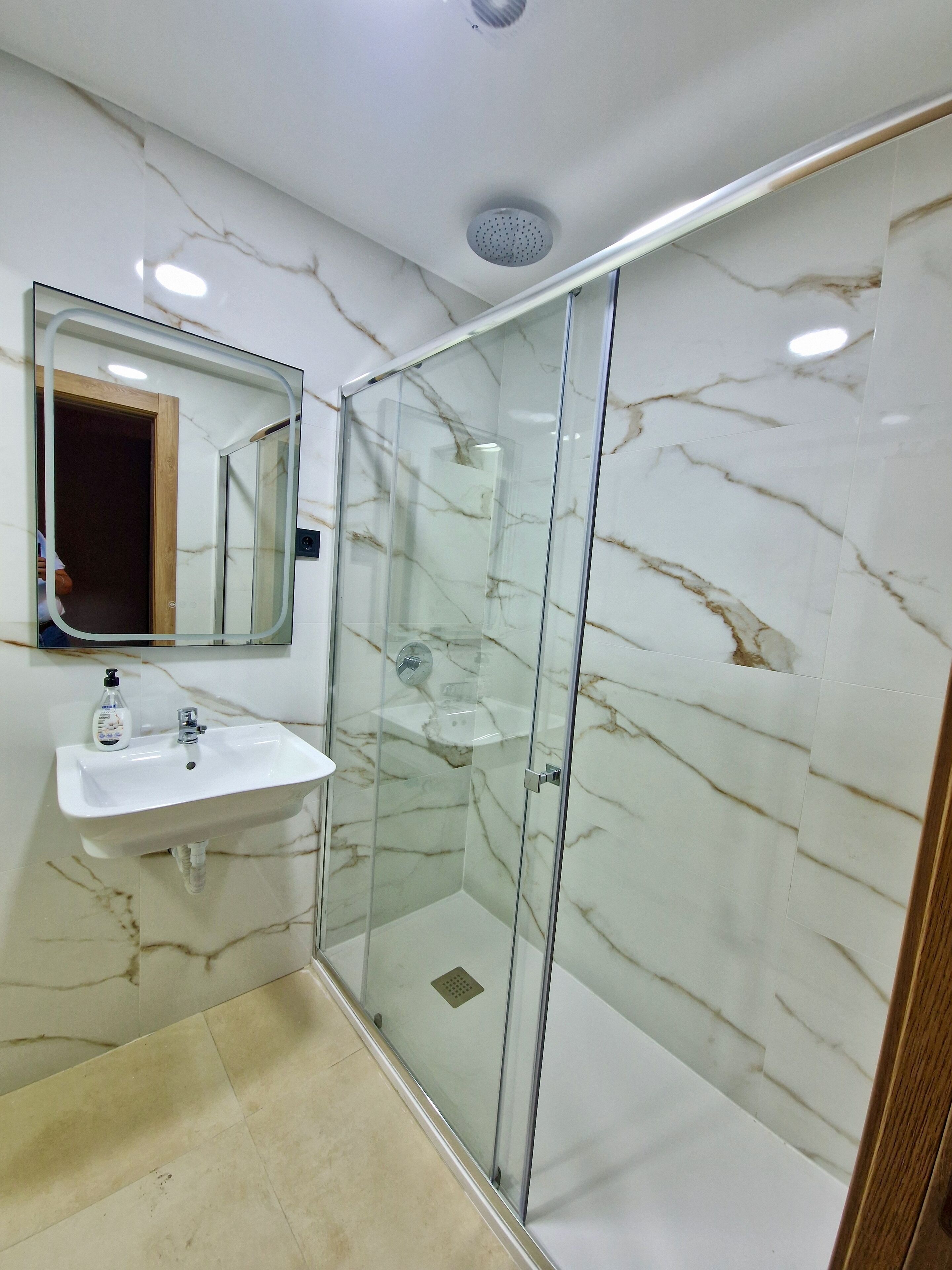 A bathroom with a glass-enclosed shower, a marble wall, a sink with a mirror, and a handheld soap dispenser.