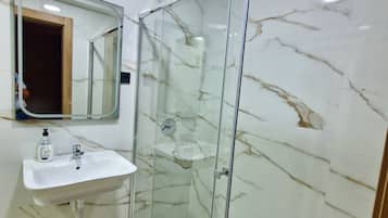 A bathroom with a glass-enclosed shower, a marble wall, a sink with a mirror, and a handheld soap dispenser.