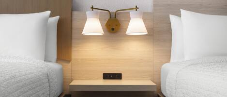 A hotel room with two beds, a wooden headboard, a nightstand with a lamp, and a power outlet.