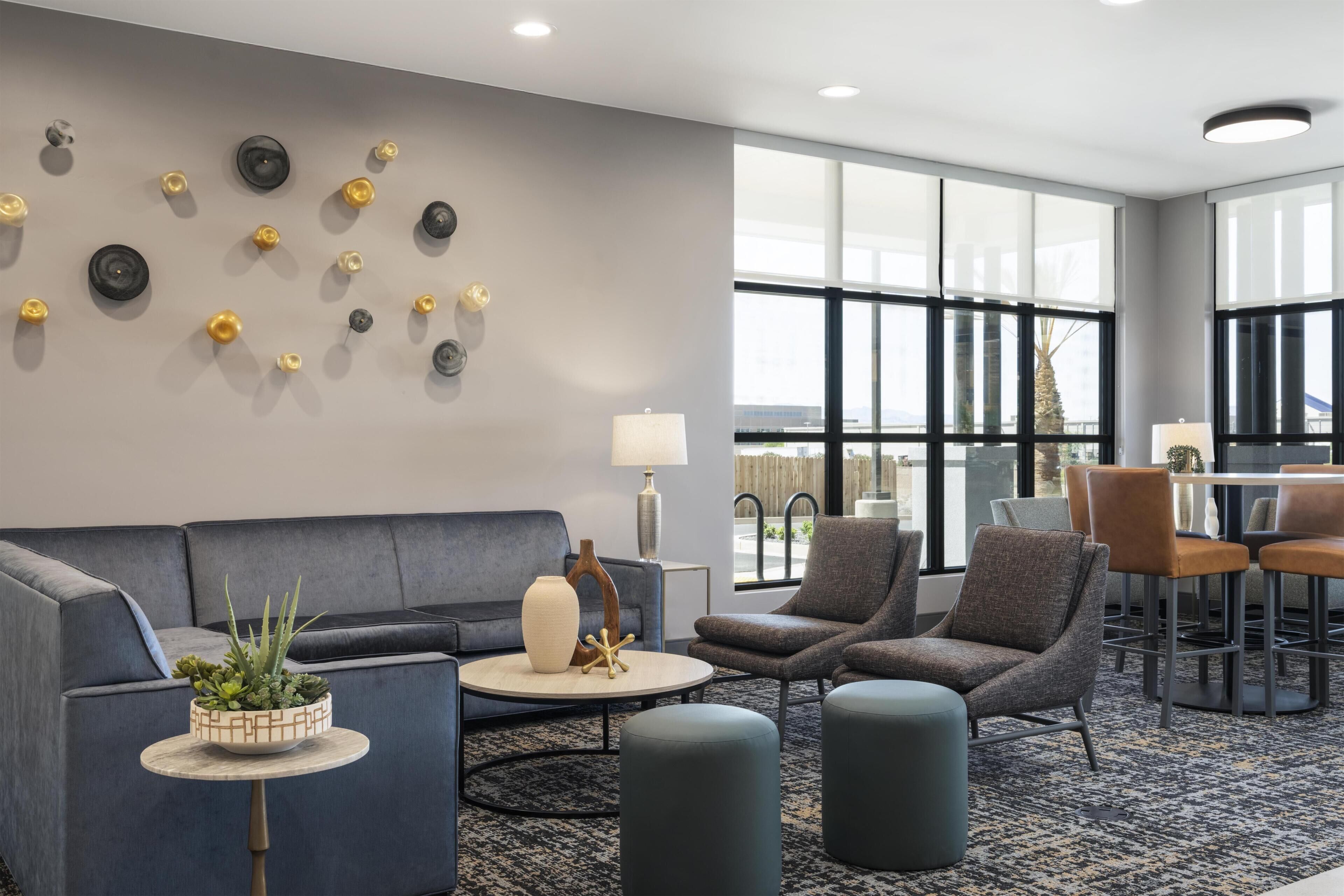 A modern hotel lobby with a grey sofa, armchairs, a coffee table, a small side table with a plant, and a large window offering a view.