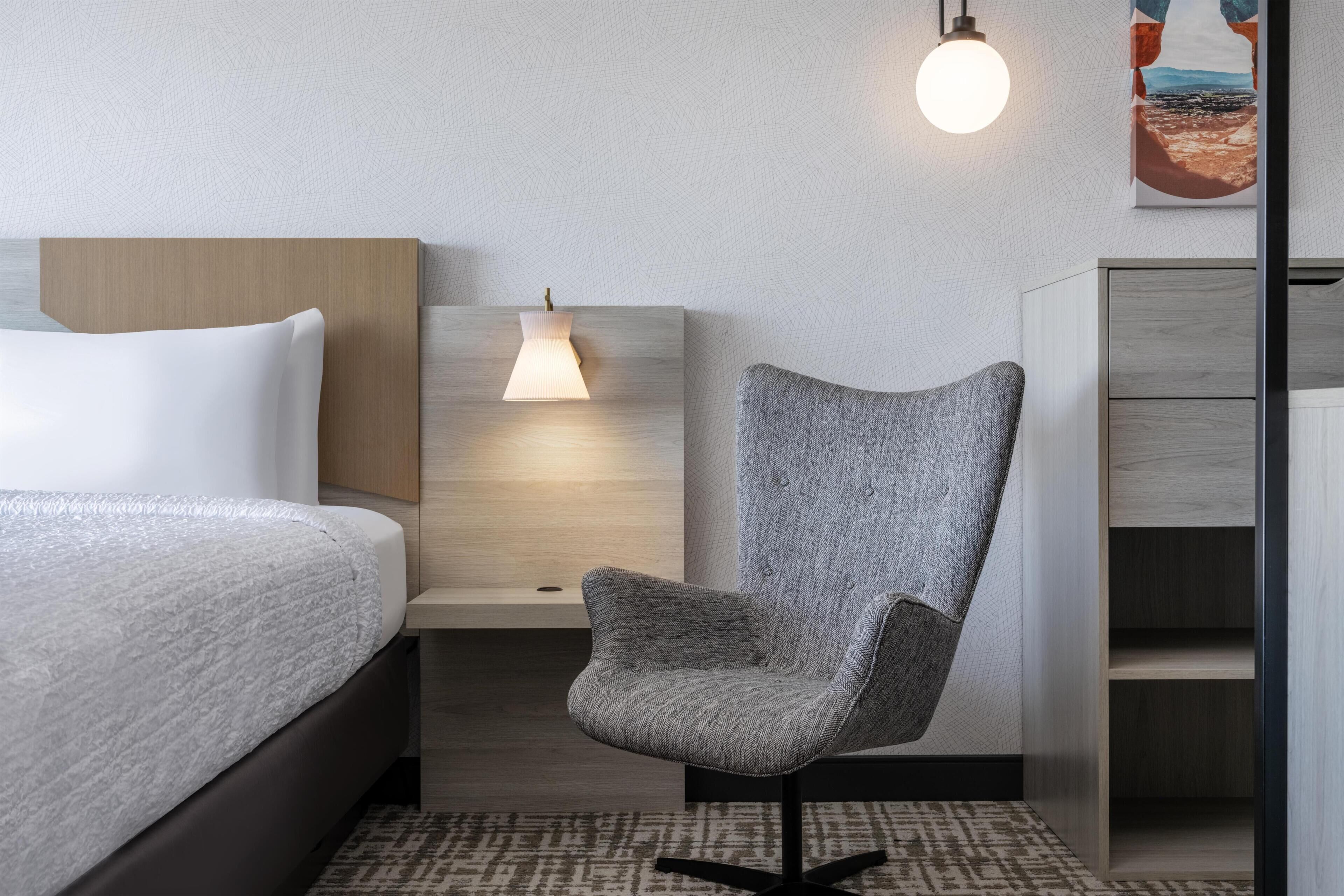 A modern hotel room with a bed, a gray upholstered chair, a bedside table with a lamp, and a wardrobe.