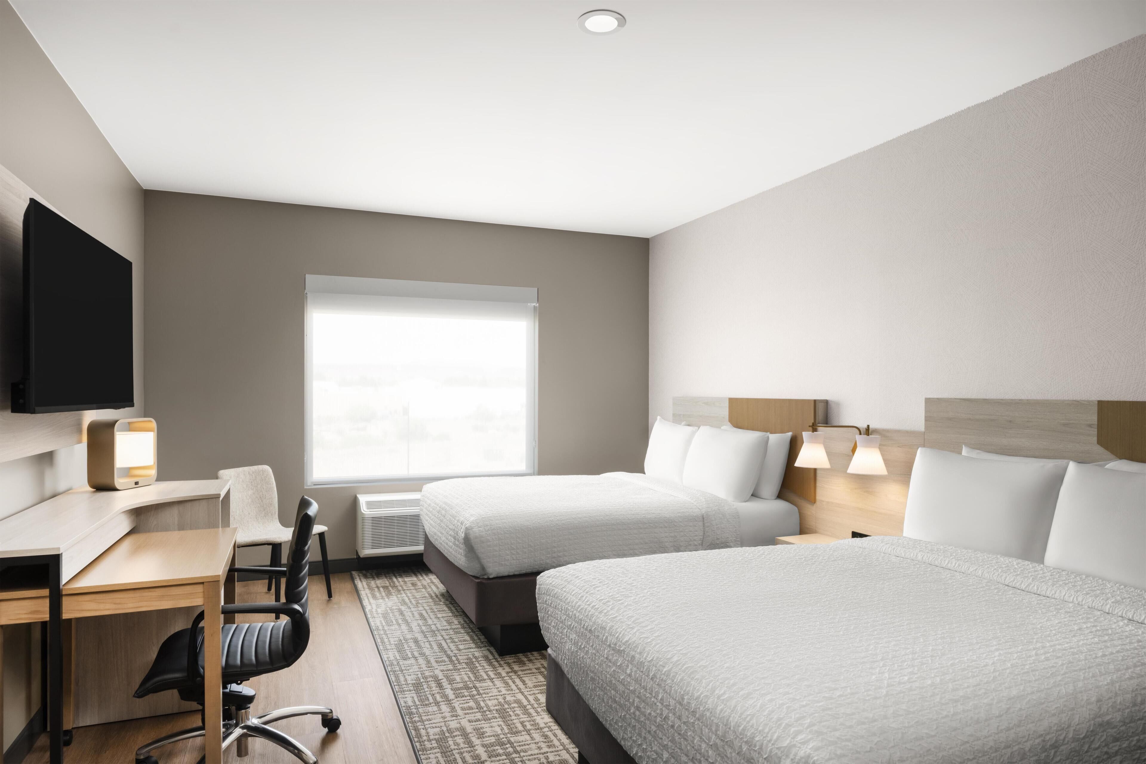 A hotel room with two beds, a desk, a chair, a TV, and a window with blinds.