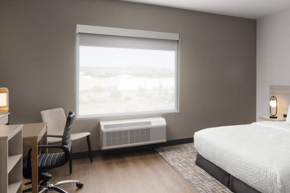 A hotel room with a bed, a desk, a chair, a window, and an air conditioning unit.