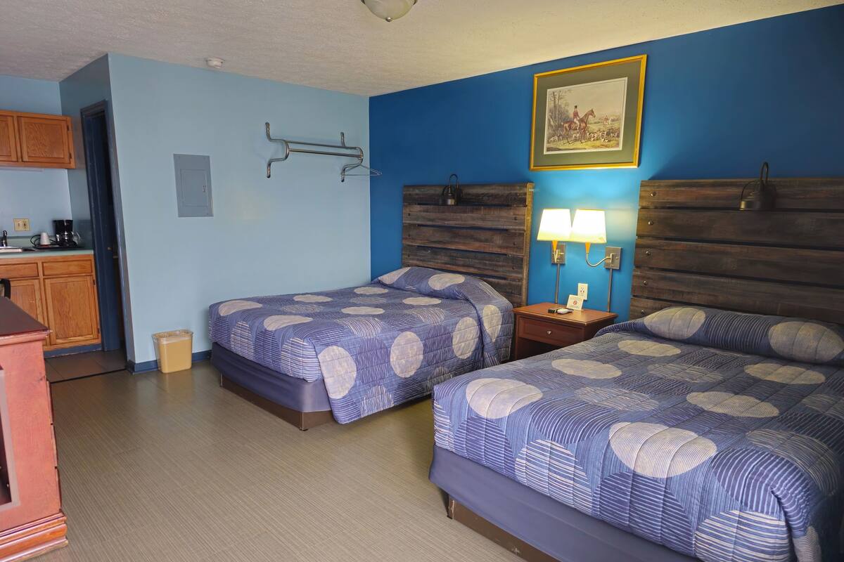 A hotel room with two beds, wooden headboards, a nightstand with a lamp, a framed picture on the wall, and a kitchenette area.