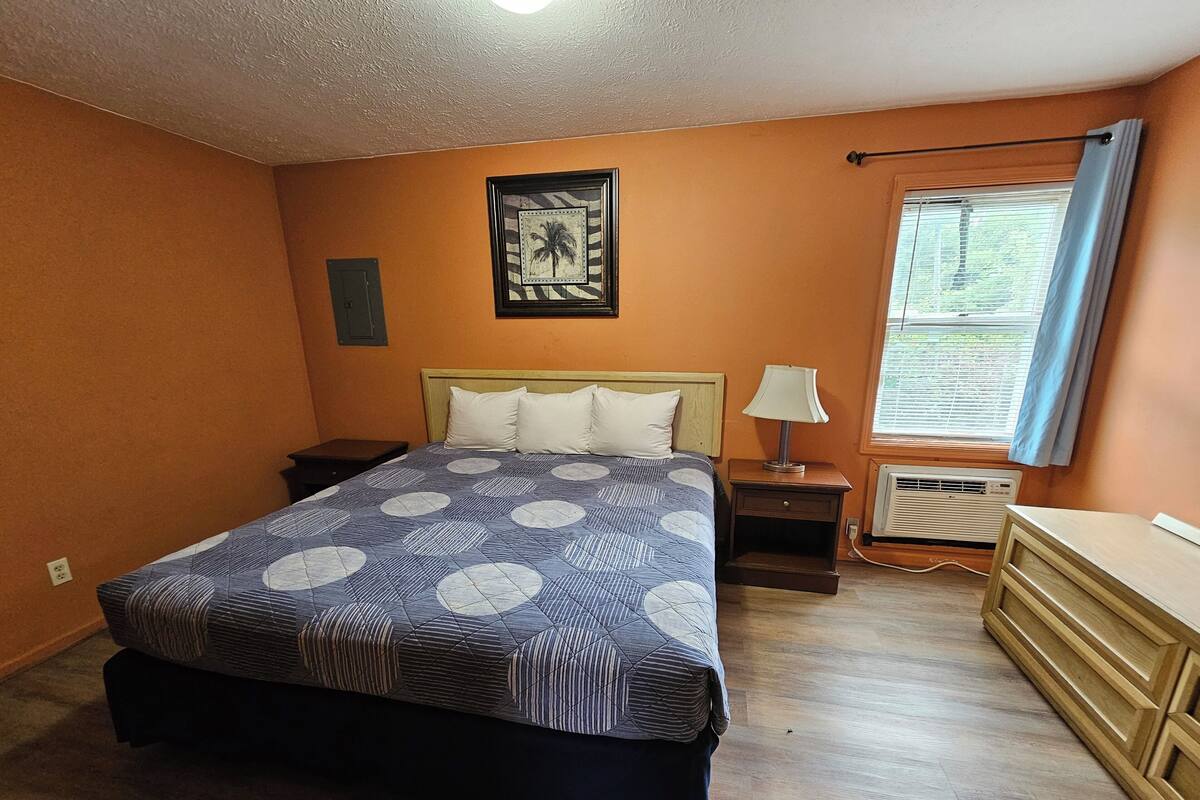 A bedroom with a bed, nightstands, a dresser, a window with blinds, and a wall-mounted air conditioner.