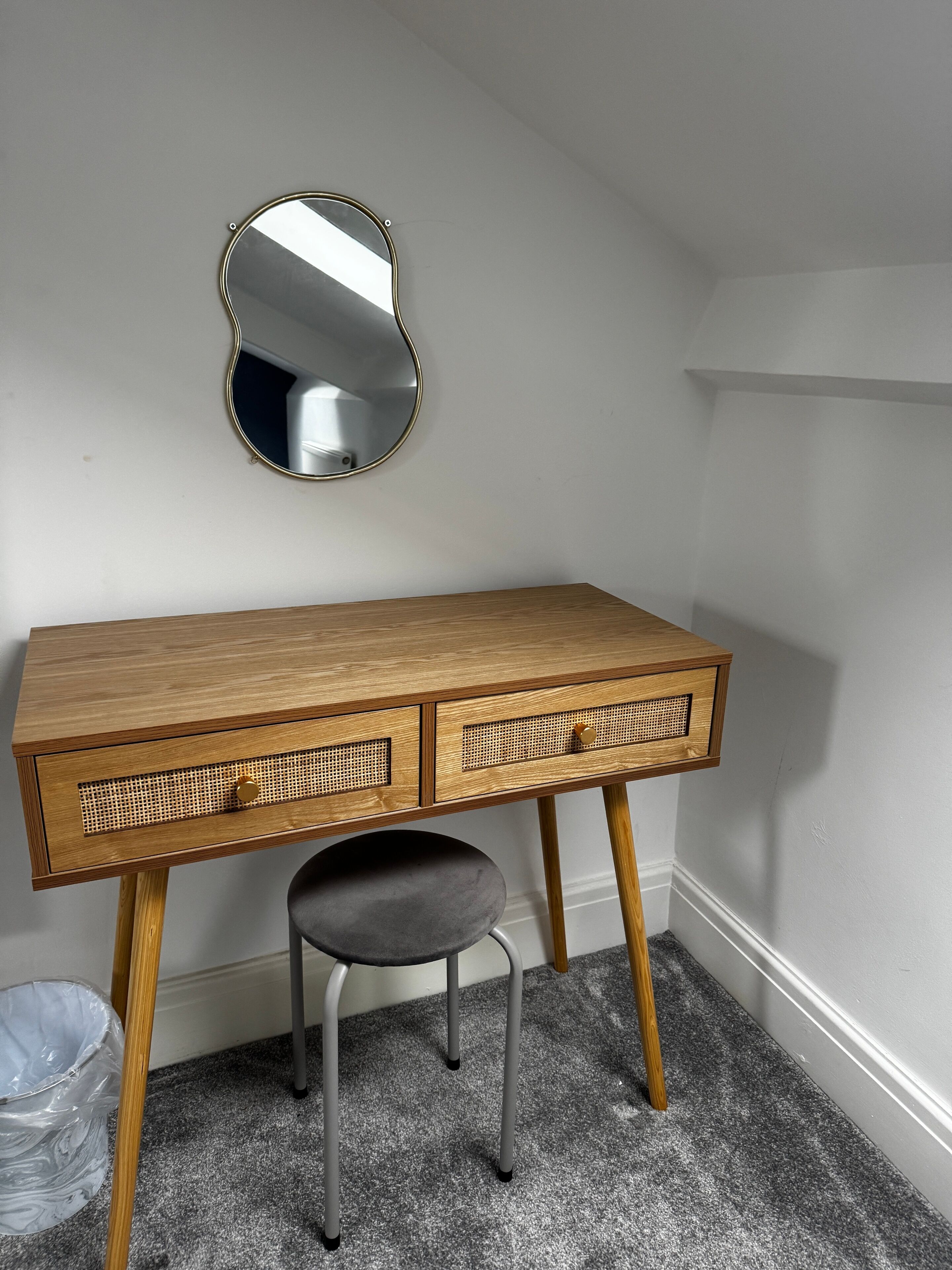 A wooden desk with two drawers and a stool, a round mirror on the wall, and a trash bin on the floor.