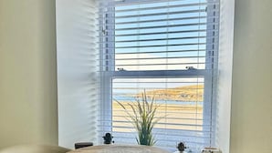 A room with a round wooden table, two chairs, a window with blinds, and a view of a beach.