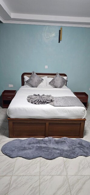 A neatly made bed with a white duvet and gray pillows, a gray fur rug on the floor, and a wooden headboard. - Juniper Park Resort (Ndaragwa)