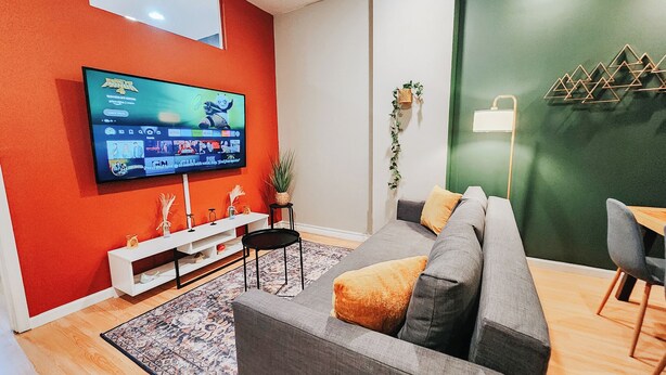 A modern living room with a flat-screen TV, a grey sofa, a small table, and a green accent wall. - Convention / 3 King Beds / Jet Shower / 65" TV (Philadelphia)