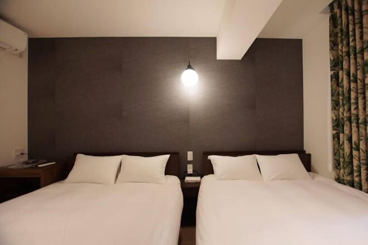 A hotel room with two beds, a nightstand with a lamp, and a large window with curtains.