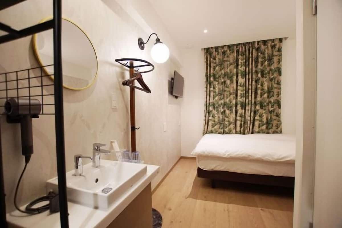 A hotel room with a bed, a sink, a mirror, and a wall-mounted lamp.