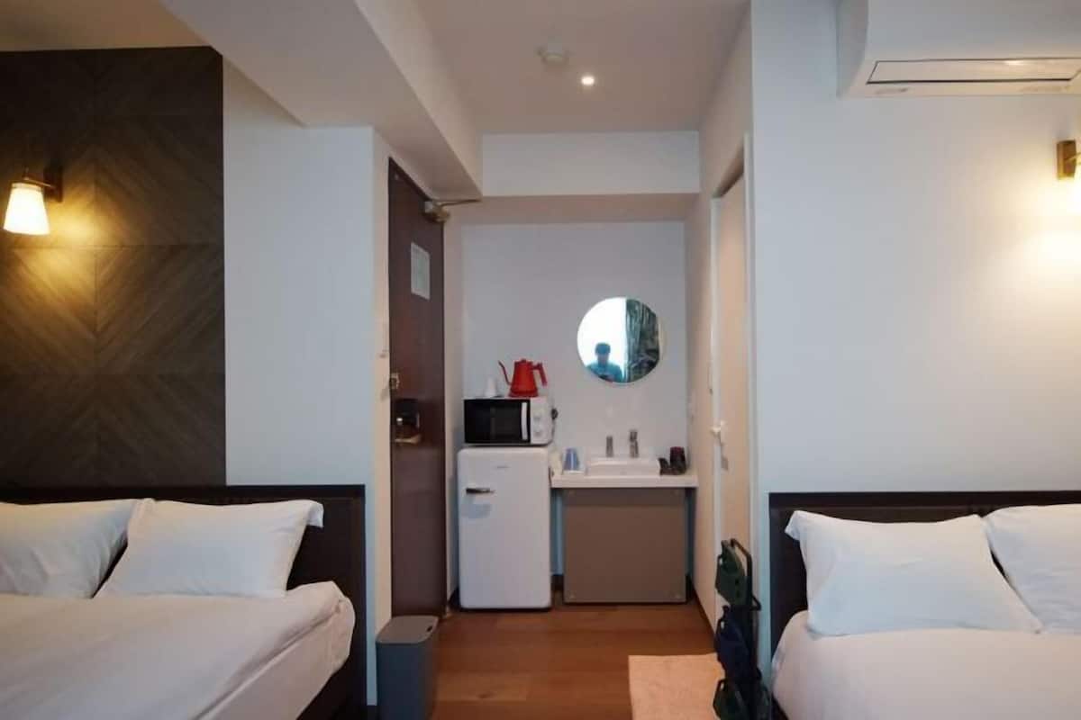 A compact hotel room with two beds, a small kitchenette, a mirror, and a ceiling light.
