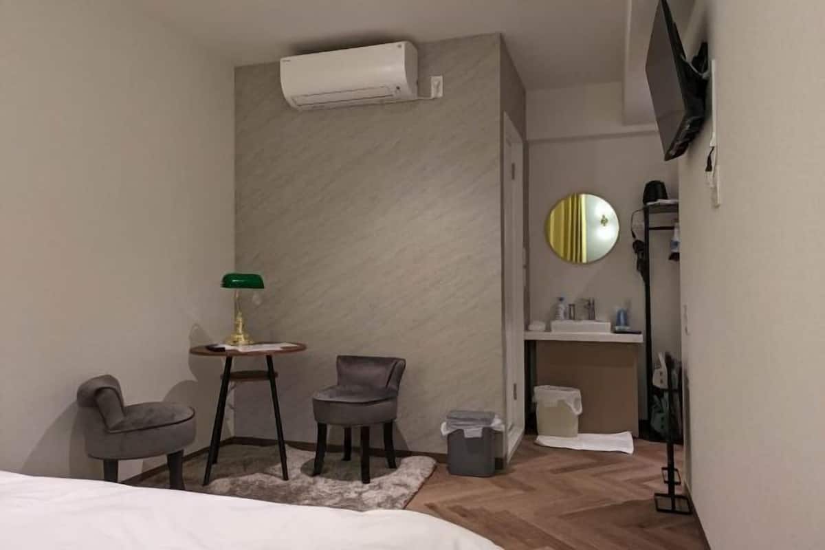 A modern hotel room with a bed, two armchairs, a small table with a lamp, an air conditioning unit, a mirror, a shelf with toiletries, and a trash bin.