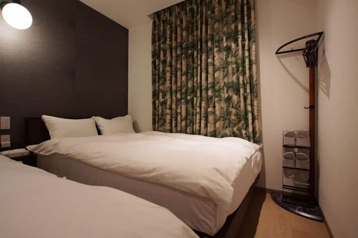 A hotel room with two beds, a dark wall, a curtain with a leaf pattern, and a shoe rack.
