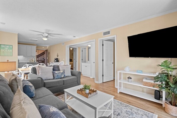 A modern living room with a grey sofa, white coffee table, and a flat-screen TV on a white stand. There is a kitchen in the background with white cabinets and a dining area. - 2-Bdrm Condo Meets Family Desires - Beachfront Resort - Full Amenities - C13 (Hilton Head Island)