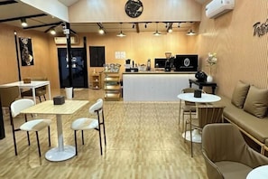A restaurant with a wooden floor, white tables, and chairs, a counter with a black and white theme, and a sofa area. - Ta Resort (Dan Sai)