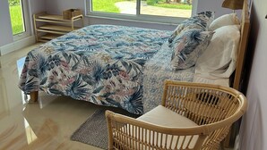Un dormitorio con cama, una silla de mimbre, una ventana que ofrece vistas a vegetación y una masa de agua.