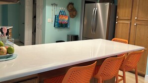 Una cocina con una isla grande, sillas anaranjadas, un refrigerador de acero inoxidable y paredes color verde azulado.
