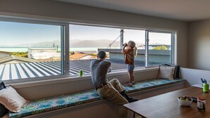 Habitación con un ventanal grande que da a un paisaje urbano y montañas. Hay un banco con cojines, una mesa con un frutero, y una persona tomando una fotografía.