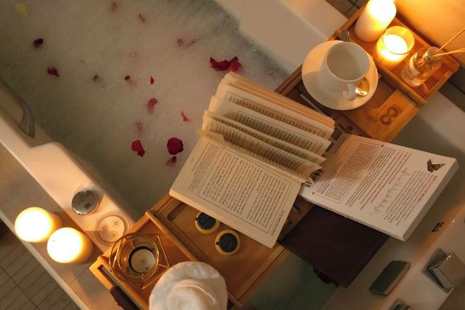 A bathtub with rose petals, a book, a cup, and a tray with candles and a candle holder.