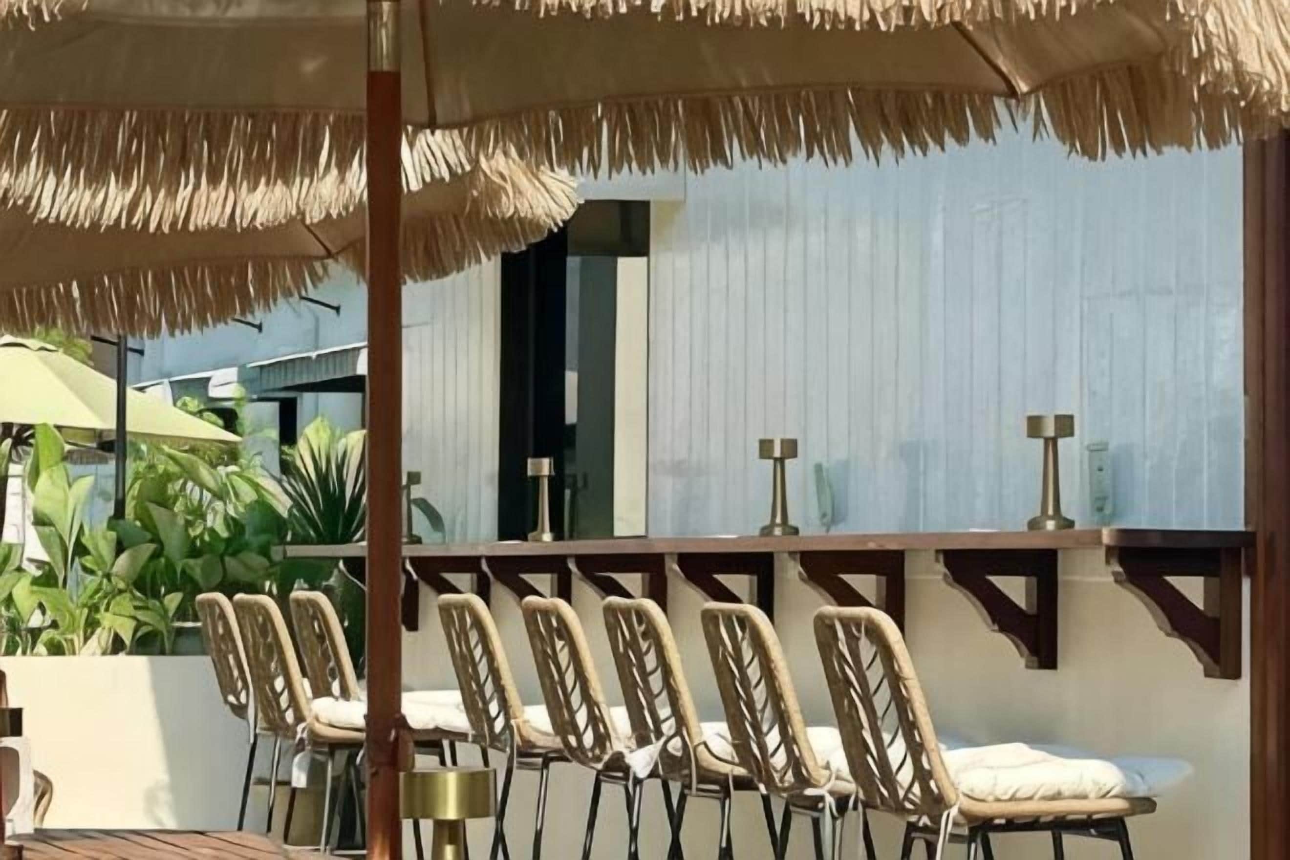 A covered outdoor seating area with chairs and tables, a thatched umbrella, and a view of a building with a window.