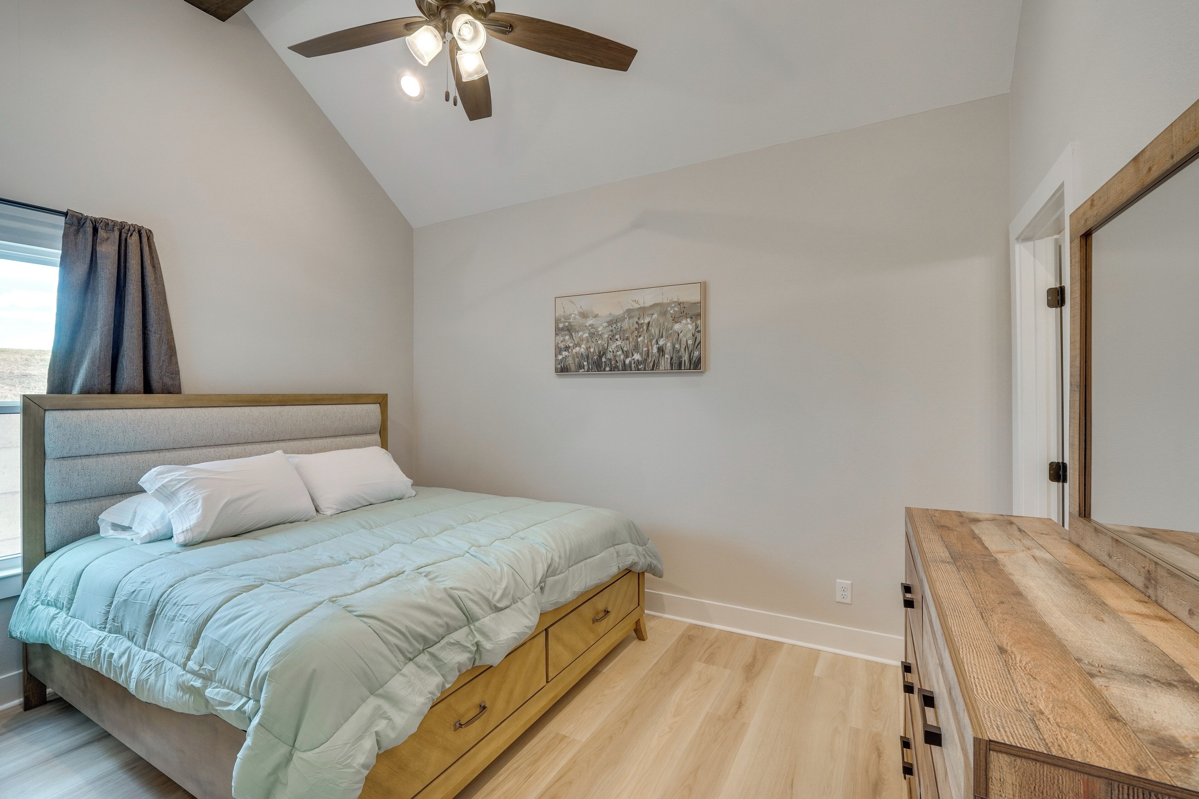 Une chambre à coucher avec un lit, une commode en bois, un ventilateur de plafond et un tableau accroché au mur.
