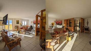 Un vaste espace de vie comprenant un grand canapé, des fauteuils et une table à manger. Un téléviseur est fixé au mur, et la pièce est dotée d’un sol carrelé.
