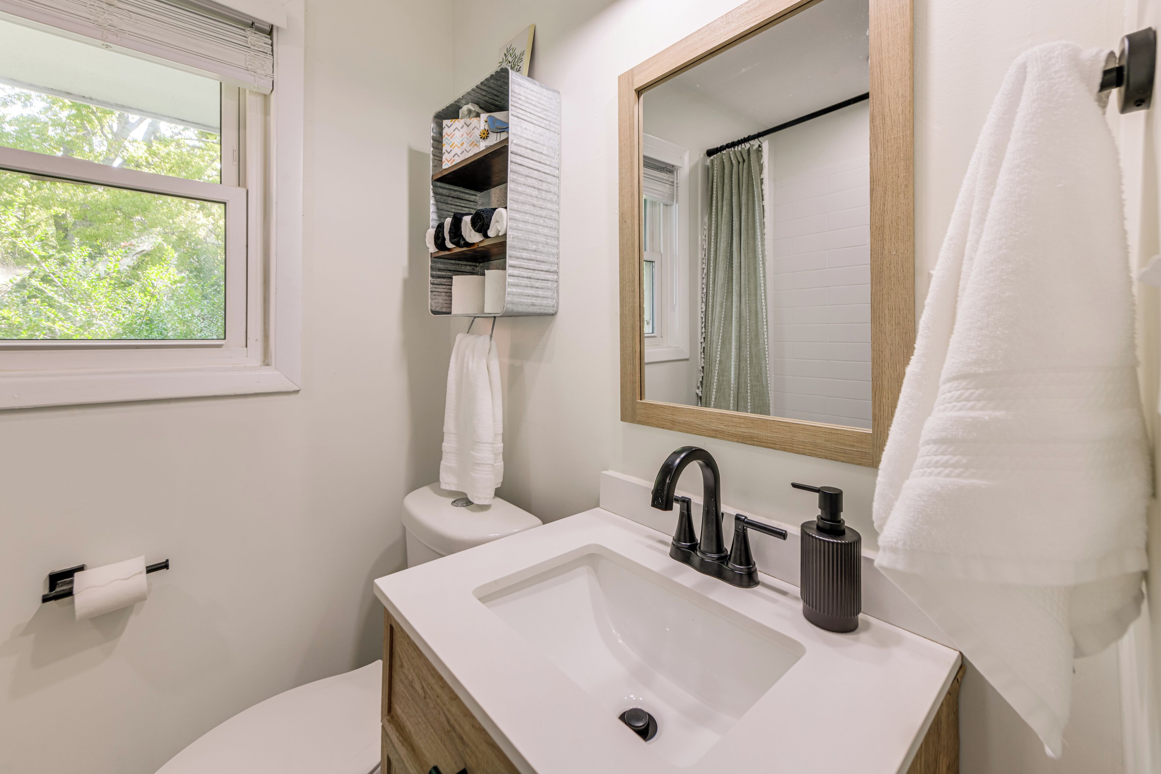 Une salle de bain moderne avec un lavabo blanc, un robinet noir et un miroir au-dessus. Une fenêtre donne sur l’extérieur, avec vue sur les arbres, et une serviette est accrochée au mur.