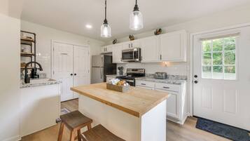 A kitchen with white cabinets, a central island, wooden stools, a microwave, a refrigerator, and hanging pendant lights.
