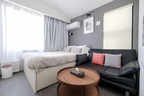 Una habitación de hotel moderna con cama, sofá, una mesa redonda de madera y un gran ventanal con cortinas.