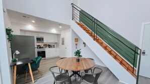 A modern kitchen with white cabinets, a dining area with a round wooden table and four chairs, and a staircase with green railings.