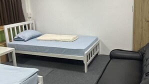 Une petite chambre avec un lit simple, une tête de lit blanche, un matelas gris, un oreiller bleu et une couverture pliée. Le sol est recouvert d’un tapis gris et un canapé en cuir noir est placé contre le mur.