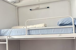 Una cama litera con colchón y almohada, un estante en la pared y una ventana con persianas.
