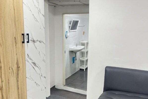 A room with a marble wall, a grey sofa, and a view of a bathroom with a sink and shelves. - JC Home Stay (Bangkok)