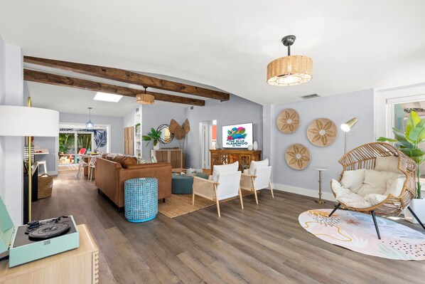 Un salon spacieux avec un canapé, des fauteuils, une table basse et un téléviseur. Le plafond est en bois, il y a une lampe et une plante.