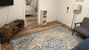 Une pièce avec un plancher en bois, un tapis à motifs, un canapé gris, une bibliothèque, une porte blanche et un téléviseur fixé au mur.
