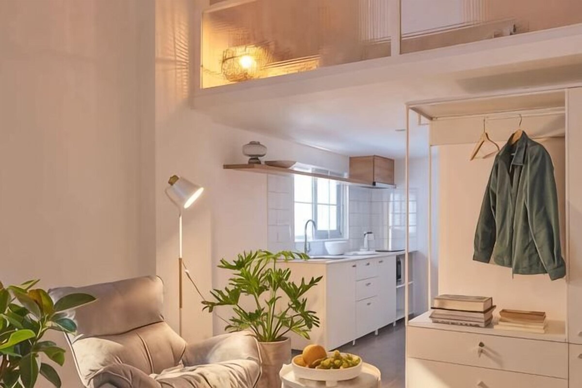 A modern living room with a sofa, a side table with fruits, a potted plant, and a kitchen in the background.