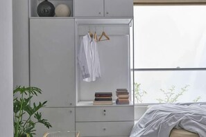 Un dormitorio con cama, un armario con ropa colgada, estantería con libros y una ventana con vistas.