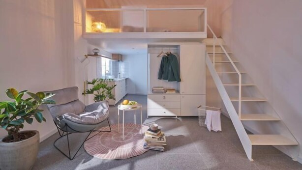 Une petite pièce comprenant un lit superposé, une chaise, une petite table avec des livres, une plante en pot et un escalier.