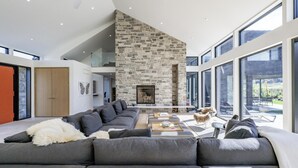 Uma sala de estar moderna com lareira de pedra, janelas amplas e um sofá modular cinza.