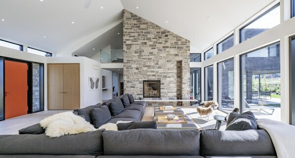 A modern living room with a stone fireplace, large windows, and a grey sectional sofa. - Luxurious new 4 bed 5 bath home (The Blue Mountains)