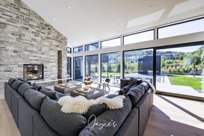 Ein modernes Wohnzimmer mit Steinwand, einem großen grauen Sofa, einem Kamin und Blick auf einen Garten durch große Fenster.