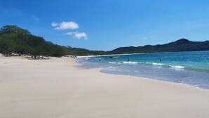 Una amplia playa de arena con olas suaves, cielo azul despejado y una colina boscosa al fondo.