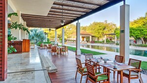 Un área de comedor al aire libre techada con mesas y sillas, una piscina y un jardín al fondo.