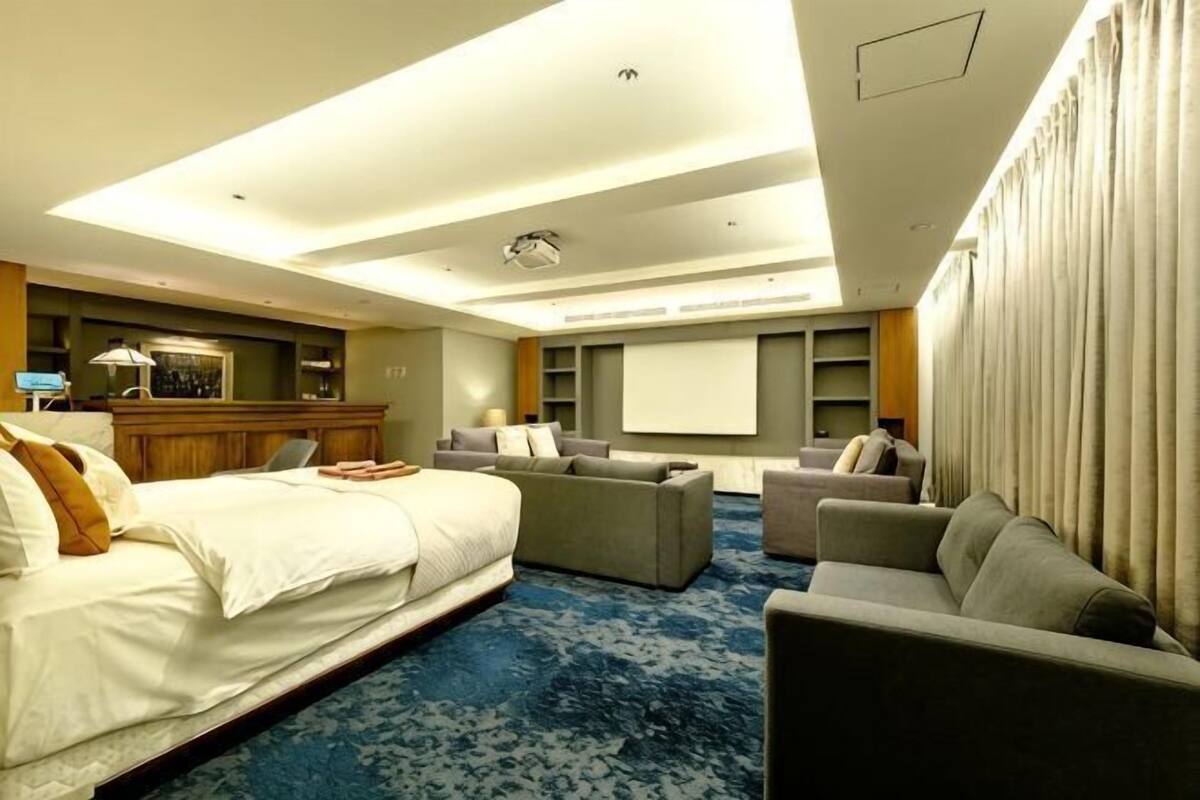 A hotel room with a large bed, a sofa, a desk with a computer, and a projector screen.