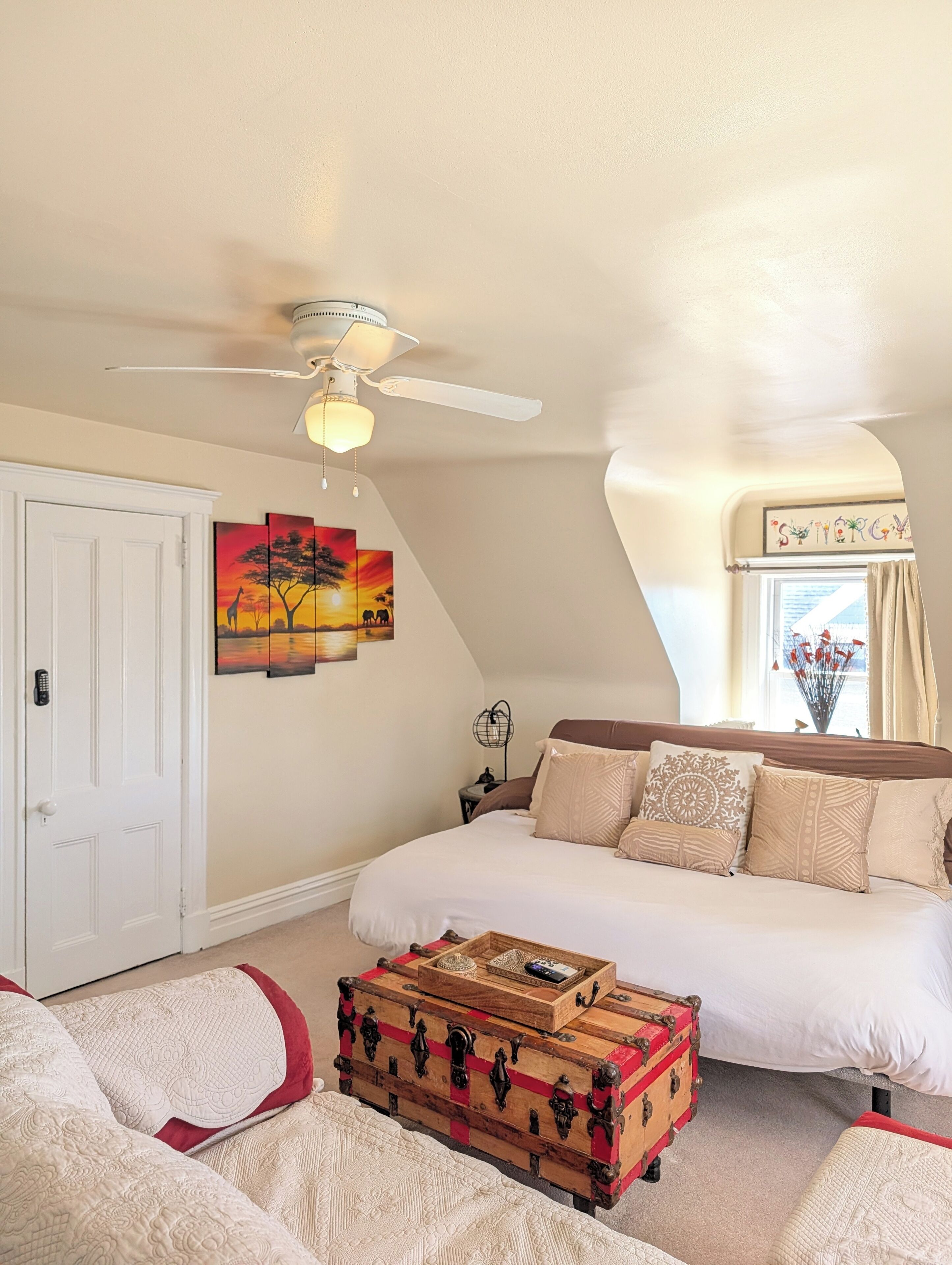 A bedroom with a bed, a ceiling fan, a wall-mounted painting, a window with curtains, and a wooden chest.