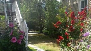 A residential garden with a pathway leading to a white porch, surrounded by flowering plants and trees. - Sunny & Comfortable: Walk To Shop, Eat & NYC Train (Montclair)